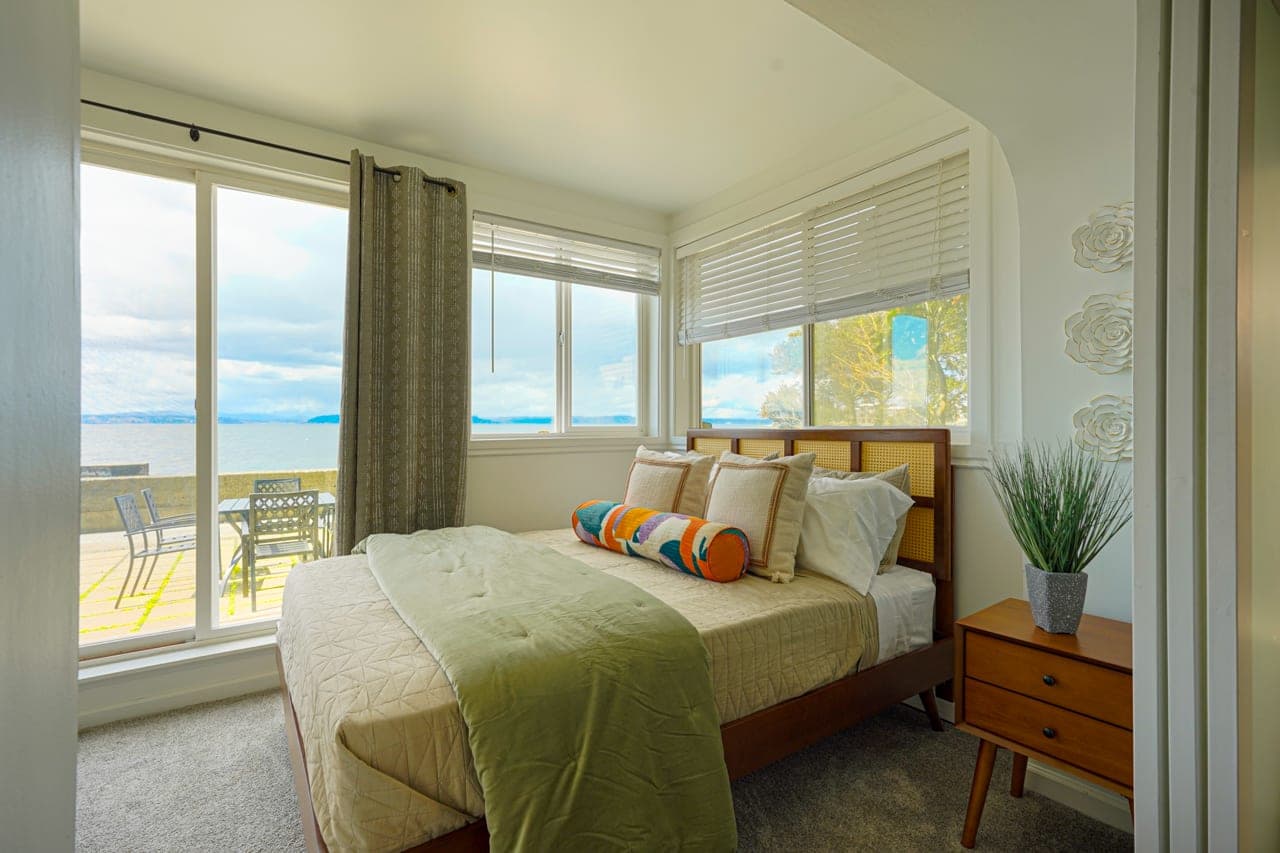This is a serene oceanfront bedroom featuring panoramic water views and mid-century modern furnishings. Wake up to the sound of waves and step directly onto a private patio for a peaceful morning by the sea.