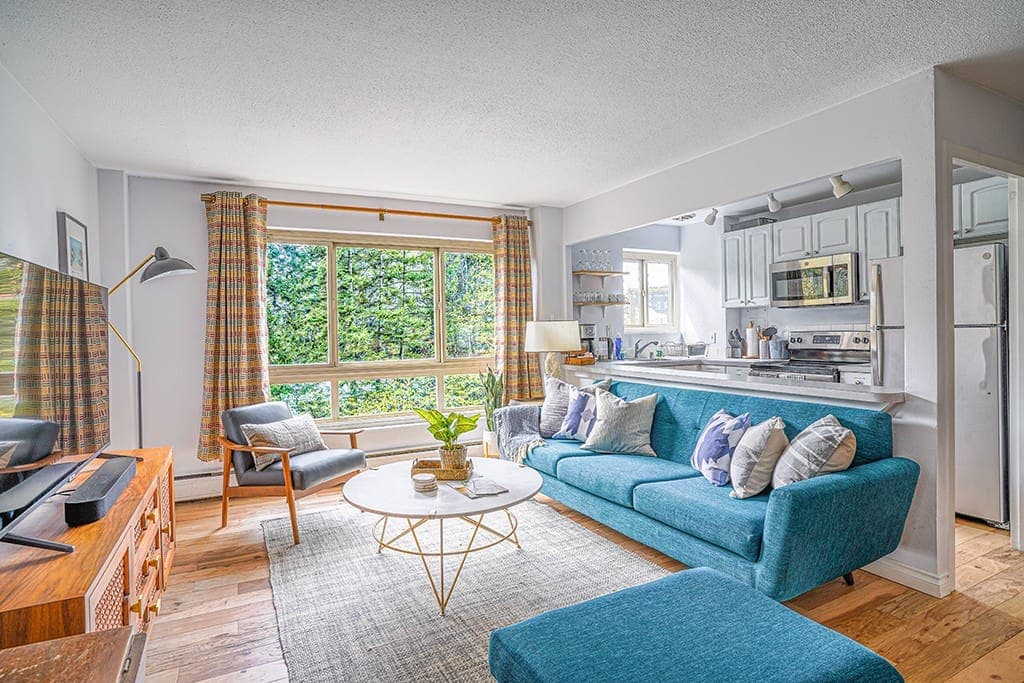 Bask in the charm of this bright and inviting open-concept living space, featuring a vibrant blue sofa, modern kitchen, and large windows with serene green views.