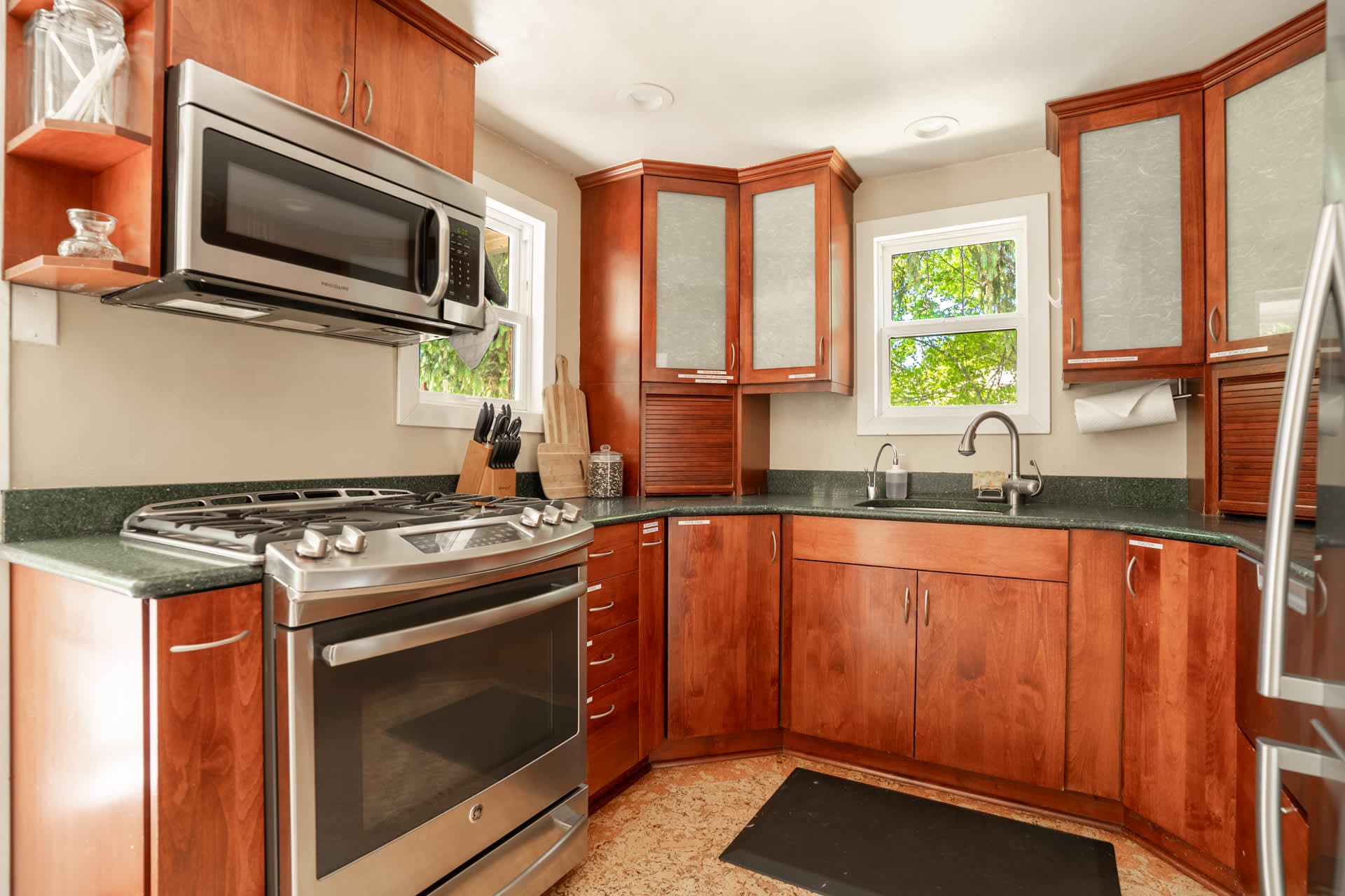 This sunlit kitchen offers a warm and functional cooking experience, with rich wood cabinetry, modern stainless steel appliances, and leafy views through the windows.